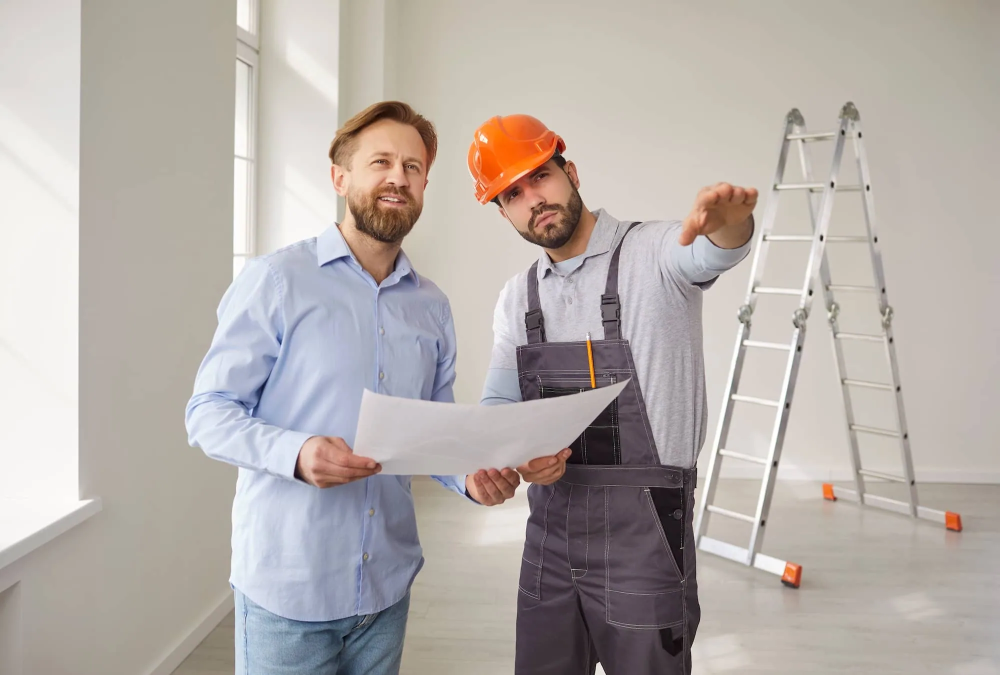 Two men reviewing blueprints in an empty room with a ladder and construction materials