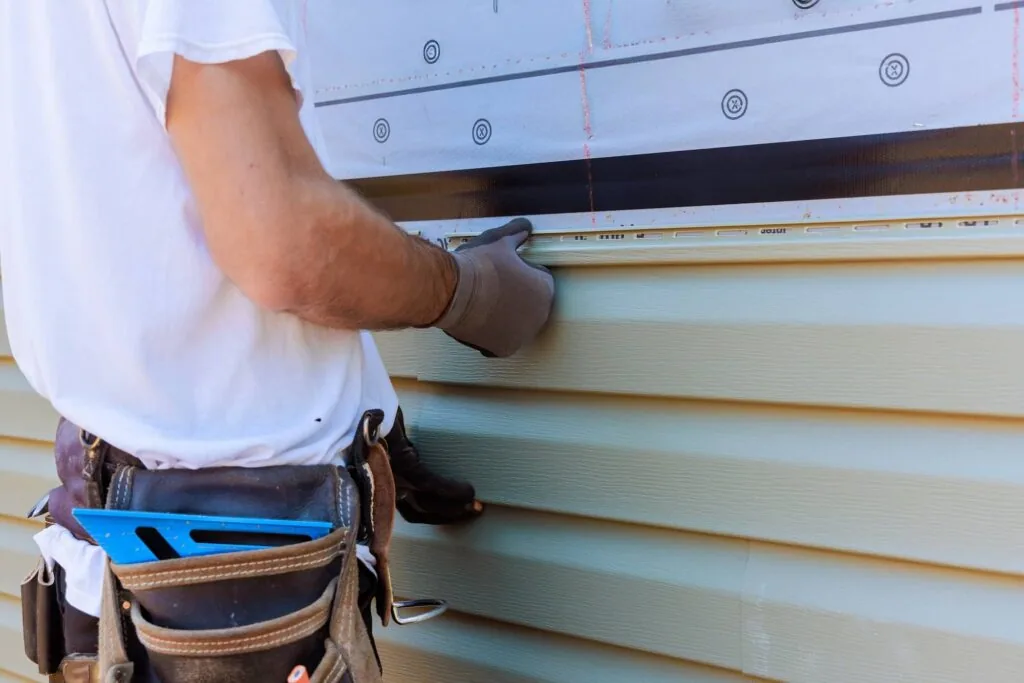 Worker in white shirt and tool belt installing or measuring light green vinyl siding on house exterior