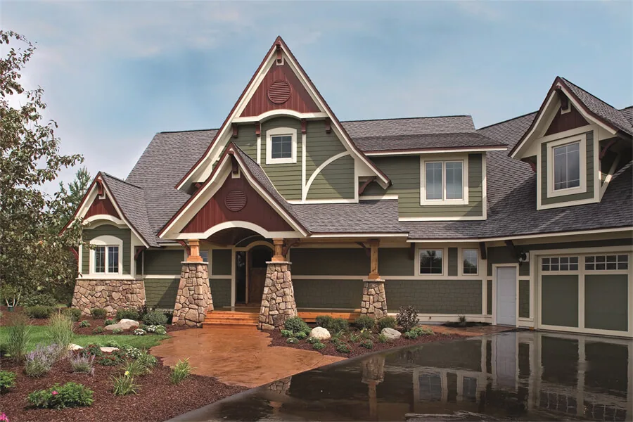 Two-story house with stone pillars, peaked roof gables, green and burgundy siding, and attached garage with landscaping.